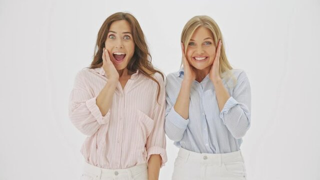 Two surprised excited women are pointing with finger to the camera and rejoicing standing isolated over white background in studio