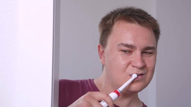 Handsome Young Man Is Up To Something, Peering Around The Corner, Brushing His Teeth With Electric Toothbrush, And Slyly Looking At Someone, Playfully Raising His Eyebrows.