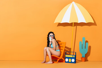 happy woman in sunglasses and swimsuit sitting on deck chair near boombox and umbrella while holding paper ice cream on orange