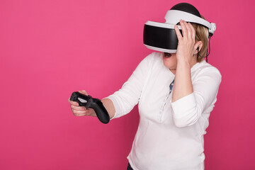 Elderly in 60s woman plays in VR helmet. She stands in white blouse on pink background and looks happy