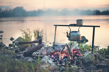Bivouac on the river at sunset,
with a water kettle hanging over the fire for tea and coffee. End of summer, goodbye to summer. A rocky shore with clumps of green grasses. The river Vistula in Poland.