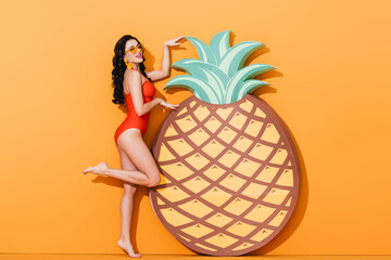 happy and barefoot woman in swimsuit and sunglasses standing near paper cut pineapple on orange, summer concept