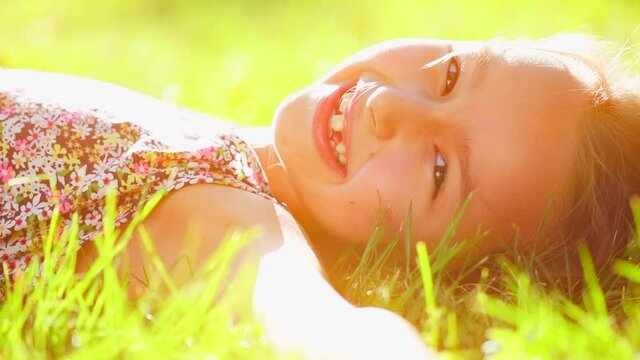 Portrait of a smiling little girl lying on green grass and dreaming. Cute six years old child enjoying nature outdoors. Healthy carefree kid playing outside in summer park on a lawn. Slow motion 4K