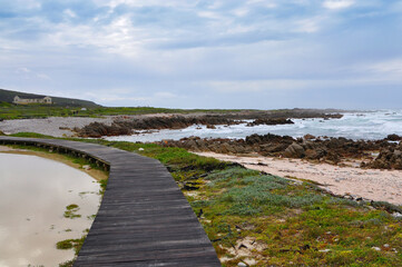 Fototapeta na wymiar Wooden boardwalk near the rocky coast and blue ocean with waves on the windy day at Cape Agulhas, the most southern point of Africa, where the Indian and Atlantic oceans meet, South Africa
