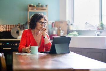 Beautiful middle-aged woman sitting in her kitchen, working on her laptop while drinking tea.
