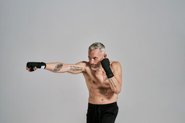 Half naked middle aged athletic man, kickboxer looking aside while boxing, practicing punches in studio over grey background