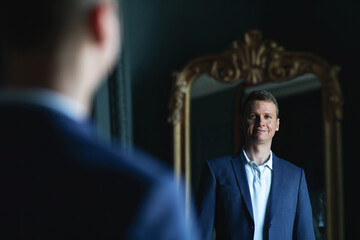 Handsome stylish man in blue jacket at home looking at mirror.
