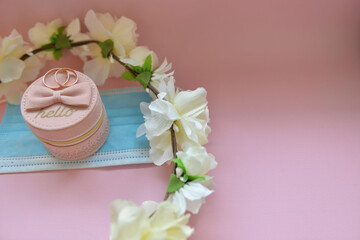 Wedding and coronavirus concept: two wedding rings, small pink box, blue medical mask (protective mask) and white flower wreath on a pink background