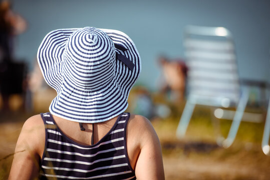 A woman in a striped hat resting by the water-view from the back.