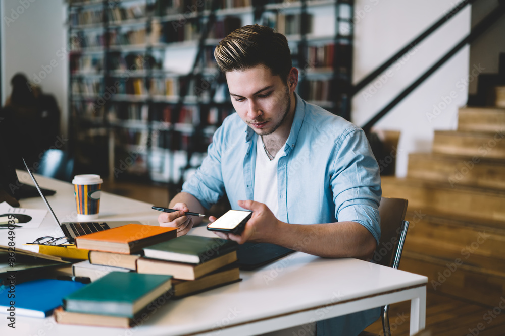 Poster pensive hipster guy using mobile phone sitting at desktop in college campus, serious millennial male