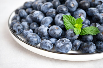 A full iron plate of ripe blueberries stands on a light gray concrete background. The bowl is decorated with fresh mint leaves. Summer seasonal berries. The concept of proper nutrition. Close-up