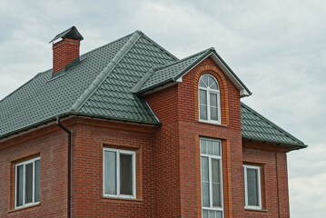 part of a red brick house with windows under a green tiled roof against a background of gray sky and clouds