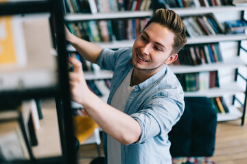 Positive hipster guy satisfied with bestsellers in book store picking new novels, smiling handsome male student spending time in university library choosing books for autodidact and education