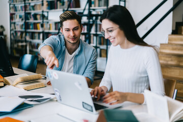 Positive male pointing on laptop computer monitor helping female installing new app for education in campus, students talking about common project making research online in library with wifi .