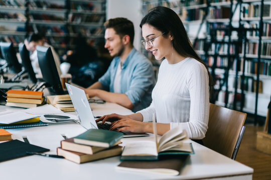 Positive young woman typing on laptop computer making research for course work in campus with wireless internet, smiling male and female students learning using modern technology in college library.