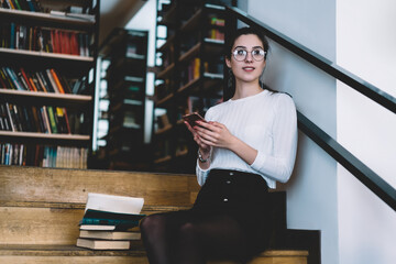 Young millennial woman in eyewear sitting on stairs in library of college connecting modern smartphone to wifi, female student using app on mobile phone for choosing literature doing homework.