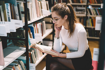 Pensive female student in eyewear for vision protection sitting near bookshelves reading...