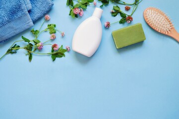 Terry towel white shampoo bottle, green soap, wooden hair brush and clover flowers on a light blue background. Flat lay natural organic cosmetic