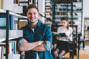 Half length portrait of cheerful hipster guy in casual outfit standing near bookshelves in college satisfied with education,happy male student looking at camera spending time in university library.