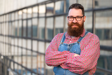 Portrait of brutal bearded stylish craftsman wearing blue overalls, checked shirt in vintage style of the mid 20th century, looking at camera, outdoors. Family workshop, old manufactory worker