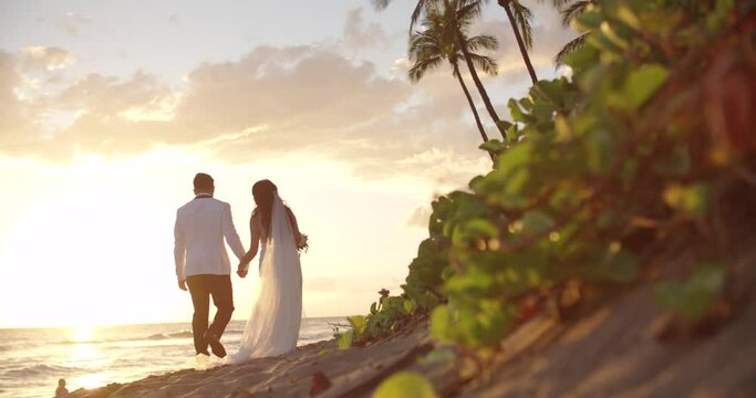 Just married Asian couple walking down tropical beach in Maui Hawaii. Wide shot with couple walking away from camera. 