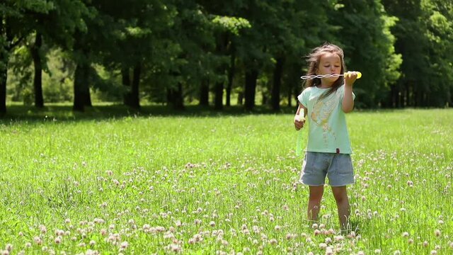 A little girl standing on a green lawn near the forest blows soap bubbles in the wind