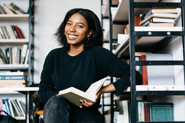 Portrait of cheerful african american girl smiling satisfied with interesting book find in college library, young dark skinned woman enjoying free time in bookstore reading literature look at camera © BullRun