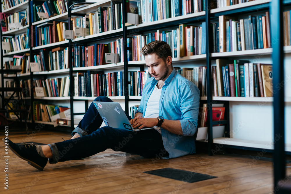 Poster pensive caucasian student in casual wear sitting on floor near big bookshelf with many literature an