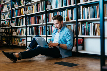 Pensive caucasian student in casual wear sitting on floor near big bookshelf with many literature...