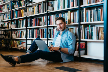 Portrait of successful young man student smiling at camera while keyboarding text for course work on laptop sitting on floor near shelf with many books.Positive hipster guy using netbook in library