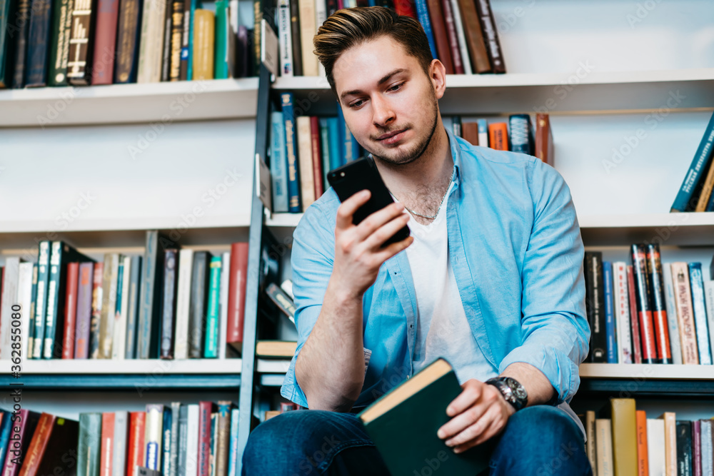 Poster hipster student reading received sms message on smartphone sitting with book in hands in public libr