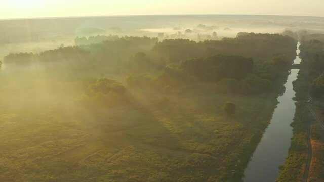 Aerial view over a valley with mist at sunrise. Slow drone flying over summer river, meadow and forest. The rays of the sun make their way through the fog. 