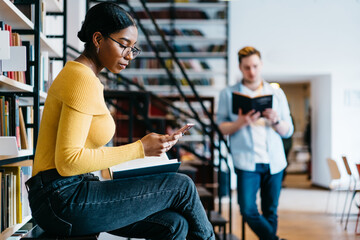 Pensive african american young woman reading incoming sms message on smartphone while preparing for exam in library.Students spending leisure time in university bookstore reading literature books