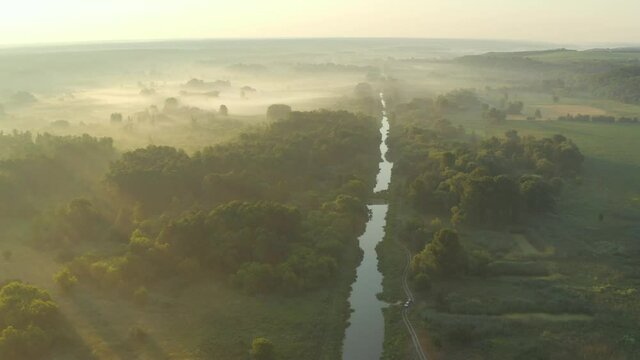 Aerial view over a valley with mist at sunrise. Slow drone flying over summer river, meadow and forest. The rays of the sun make their way through the fog. 