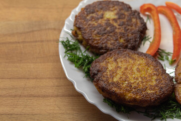 Two potato cutlets lie on a white plate along with dill and red Bulgarian pepper