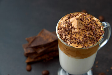 Glass of trendy fluffy creamy dalgona or whipped instant coffee with milk, coffee beans and chocolate on the dark background. New popular food and drink trend concept. Copy space.