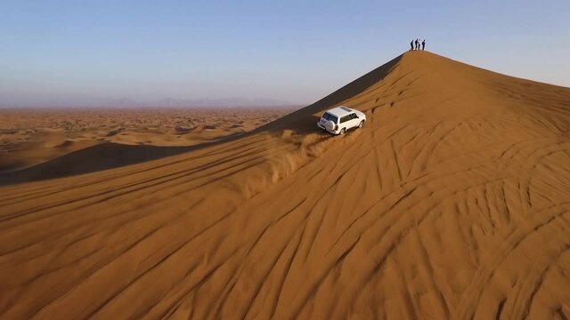 4x4 SUV driving on the edge of big sand dune towards group of people at the top, drone pulling in revealing desert landscape
