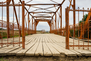 Ancient village bridge over the river. The metal frame of the Red Bridge. Wooden boards of the old bridge. Historical monument. Middle Asia. Kazakhstan. Country road. Green grass and sky