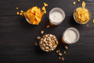 Nachos, chips, pistachios in glass bowls and scattered on wooden table, near are beer with foam in glasses