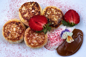 Cottage cheese pancakes, syrniki with fresh berries for breakfast on white plate, wooden table background. Russian sweet pancakes with stawberries and flowers and caramel