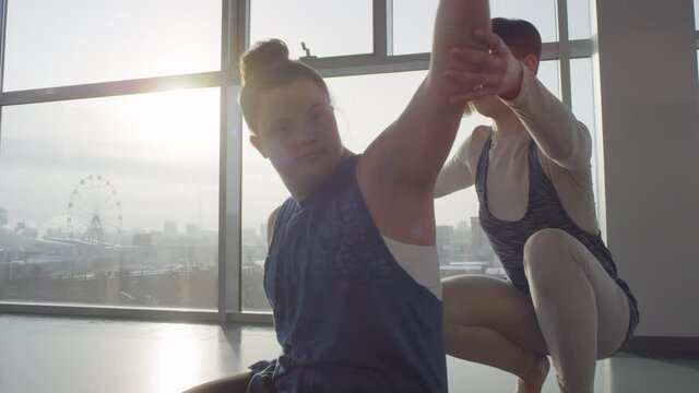Professional yoga trainer with short haircut helping disabled woman with down syndrome to stretch her arms, handheld medium shot