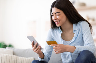 Fototapeta premium Online Payment. Smiling Asian Woman Using Smartphone And Credit Card At Home