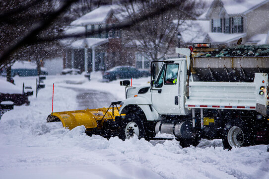 View of white and yellow city snow plow truck driving away while removing fresh deep snowfall in winter in suburban area to make roads safe for drivers