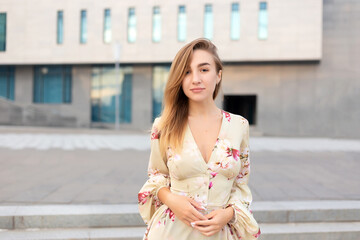 Fototapeta premium Portrait of a beautiful young girl on a background of a blue business center dressed in a summer beige dress