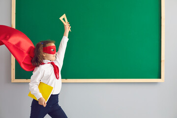 Back school. Cute brave girl in superhero costume near chalkboard at classroom. Kid wearing hero...