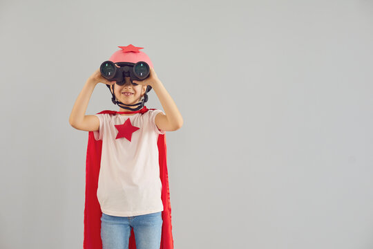 Child wearing superhero costume and looking into binoculars on grey background. Cute girl pretending to be superwoman