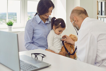 Family doctor. Mature doctor listening to little girl heartbeat in clinic. Mother with child on visit to pediatrician in hospital