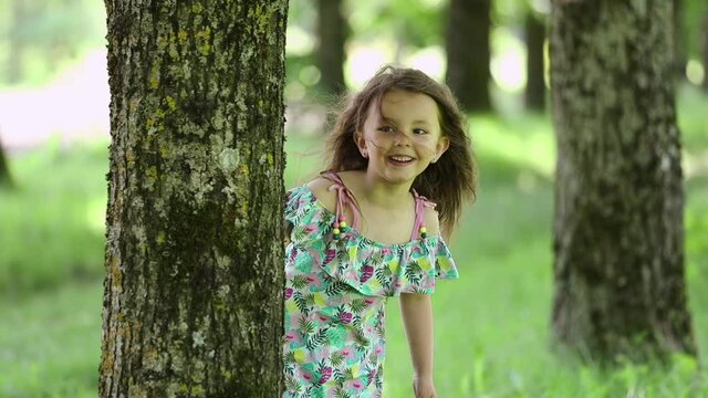 Little girl looks out from behind a tree trunk and runs away along a shady alley