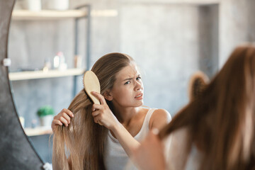 Naklejka premium Frustrated young woman trying to brush her tangled hair in front of mirror in bathroom