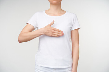 Horizontal photo of young female dressed in white t-shirt keeping raised palm on her chest while using sign language, isolated over white background